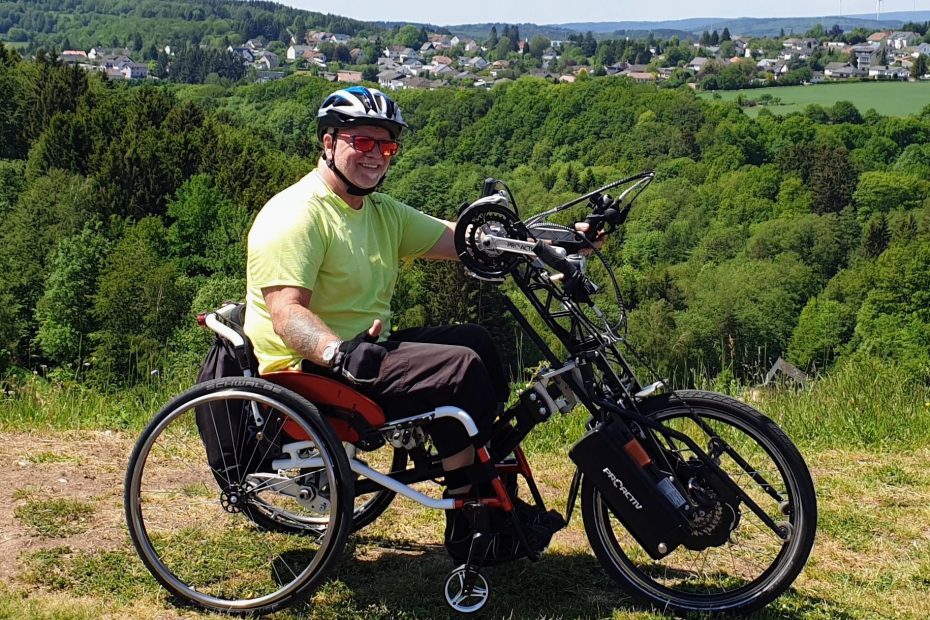 Der Behindertenbeauftragte Hartmut Heinke auf einem Adaptivbike mit Rollstuhl in der Natur. Er zeigt den Daumen nach oben und lächelt