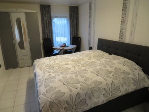 Bild von einem Schlafzimmer mit einem Doppelbett, einem dreitürigen Schrank, einem Fenster, ein Tisch mit zwei Stühlen.