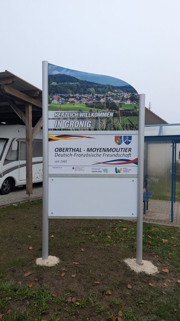 Ortseingangsschild von Gronig, weißes Schild mit Foto des Blicks über Gronig ; Herzlich Willkommen in Gronig, Schild, das die deutsch-französische Freundschaft zwischen den Partnergemeinden Oberthal und Moyenmoutier beschreibt. Weiteres weißes Schild.