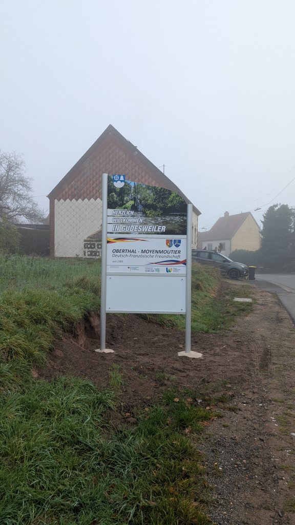 Ortseingangsschild von Güdesweiler, weißes Schild mit Foto eines Weihers ; Herzlich Willkommen in Güdesweiler, Schild, das die deutsch-französische Freundschaft zwischen den Partnergemeinden Oberthal und Moyenmoutier beschreibt. Weiteres weißes Schild.