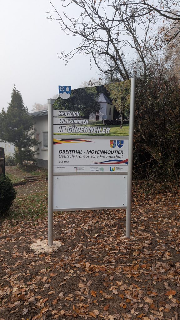 Ortseingangsschild von Güdesweiler, weißes Schild mit Foto der Valentinskapelle ; Herzlich Willkommen in Güdesweiler, Schild, das die deutsch-französische Freundschaft zwischen den Partnergemeinden Oberthal und Moyenmoutier beschreibt. Weiteres weißes Schild.