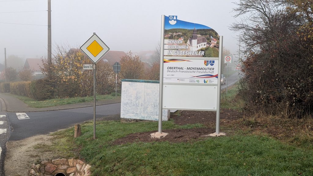Ortseingangsschild von Güdesweiler, weißes Schild mit Foto der Güdesweiler Kirche; Herzlich Willkommen in Güdesweiler, Schild, das die deutsch-französische Freundschaft zwischen den Partnergemeinden Oberthal und Moyenmoutier beschreibt. Weiteres weißes Schild.
