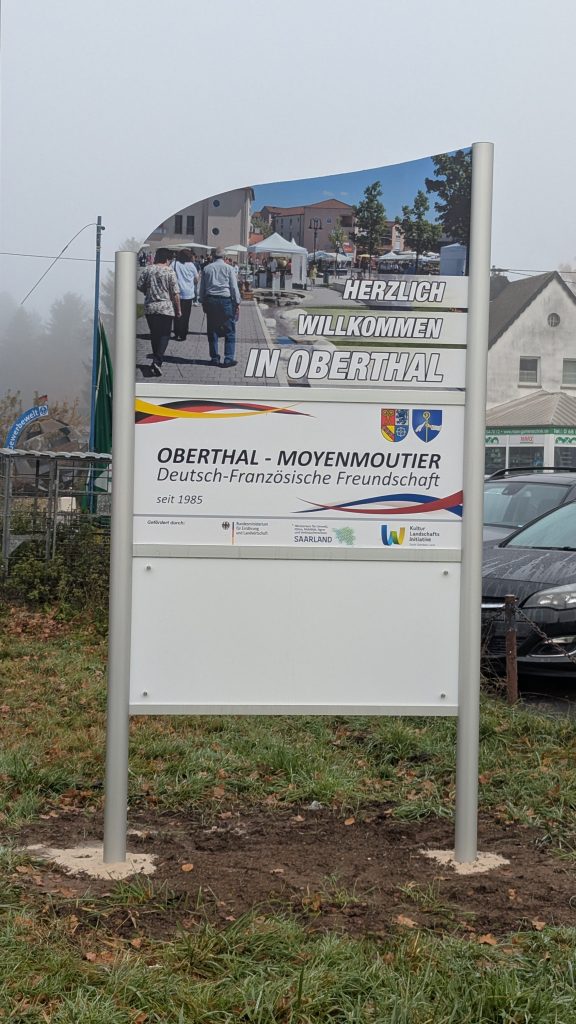 Ortseingangsschild von Oberthal, weißes Schild mit Bild vom Töpfermarkt; Herzlich Willkommen in Oberthal, Schild, das die deutsch-französische Freundschaft zwischen den Partnergemeinden Oberthal und Moyenmoutier beschreibt. Weiteres weißes Schild.