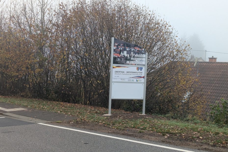 Ortseingangsschild von Oberthal, weißes Schild mit Bild von der Kirche; Herzlich Willkommen in Oberthal, Schild, das die deutsch-französische Freundschaft zwischen den Partnergemeinden Oberthal und Moyenmoutier beschreibt. Weiteres weißes Schild.