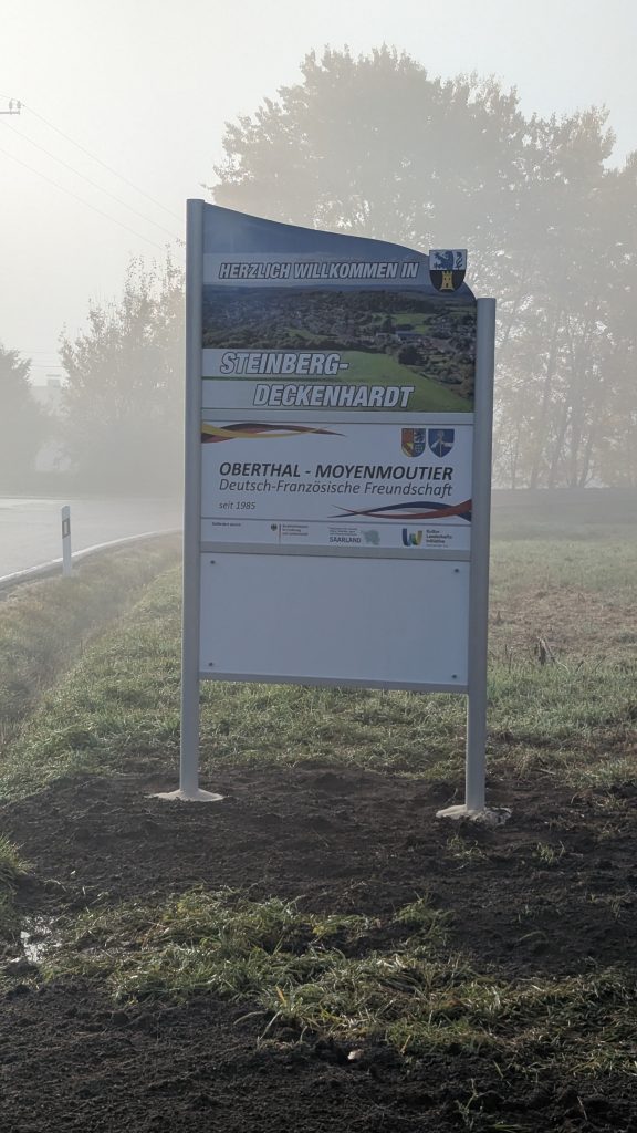 Ortseingangsschild von Steinberg-Deckenhardt, weißes Schild mit Foto mit Blick über Steinberg-Deckenhardt; Herzlich Willkommen in Steinberg-Deckenhardt, Schild, das die deutsch-französische Freundschaft zwischen den Partnergemeinden Oberthal und Moyenmoutier beschreibt. Weiteres weißes Schild.